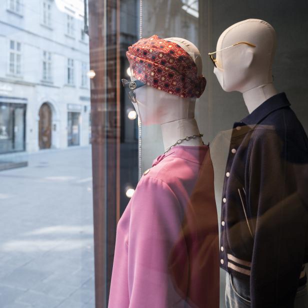Zwei Schaufensterpuppen mit Sonnenbrillen und modischer Kleidung blicken durch ein Fenster auf eine ruhige Straße.