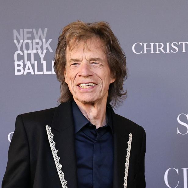 Mick Jagger lächelt vor einem grauen Hintergrund mit Logos.