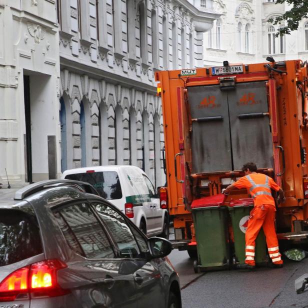 Ein Müllwerker in orangefarbener Arbeitskleidung leert Mülltonnen in ein orangefarbenes Müllfahrzeug auf einer Straße.
