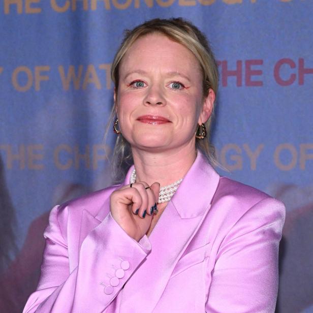 Thora Birch mit blonden Haaren trägt einen rosa Blazer und eine Perlenkette vor einem blauen Hintergrund.