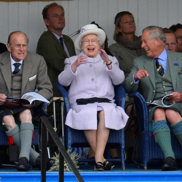 Queen Elizabeth II. und Charles lachen, während sie in traditioneller schottischer Kleidung auf Stühlen sitzen. Prinz Philip ist auch dabei.