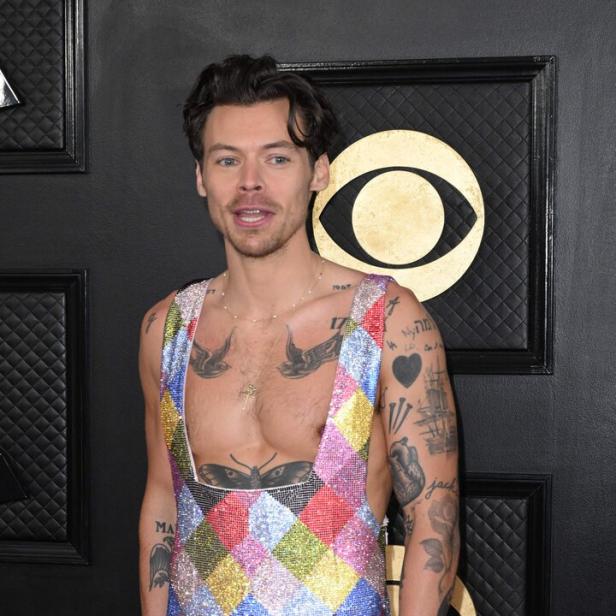 Harry Styles trägt einen bunten, rautenförmigen Overall und zeigt seine Tattoos.