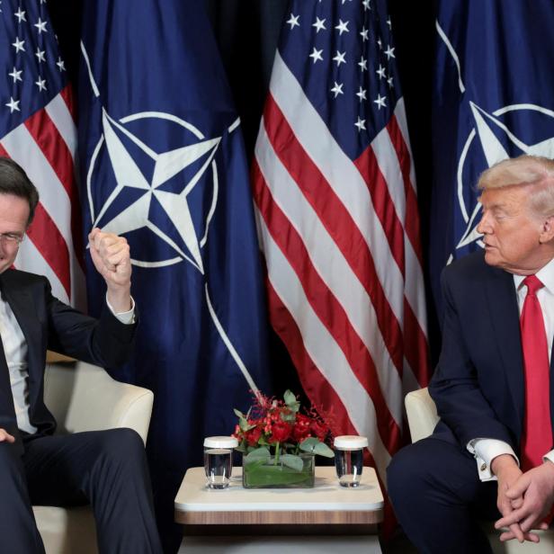 Mark Rutte gestikuliert neben Donald Trump vor US- und NATO-Flaggen.