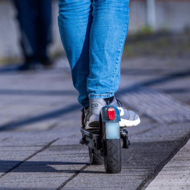 Person in blauer Jeans fährt auf einem E-Scooter auf einem gepflasterten Gehweg.