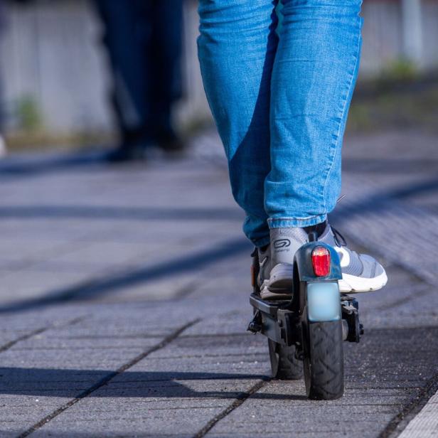 Person in blauer Jeans fährt auf einem E-Scooter auf einem gepflasterten Gehweg.
