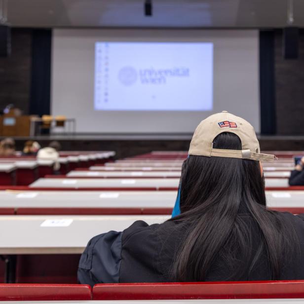 Eine Studentin mit Kappe sitzt in einem Hörsaal der Universität Wien vor einer Leinwand.