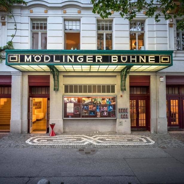 Eingang eines Theaters mit beleuchtetem Vordach, großen Fenstern und Plakaten an der Wand.