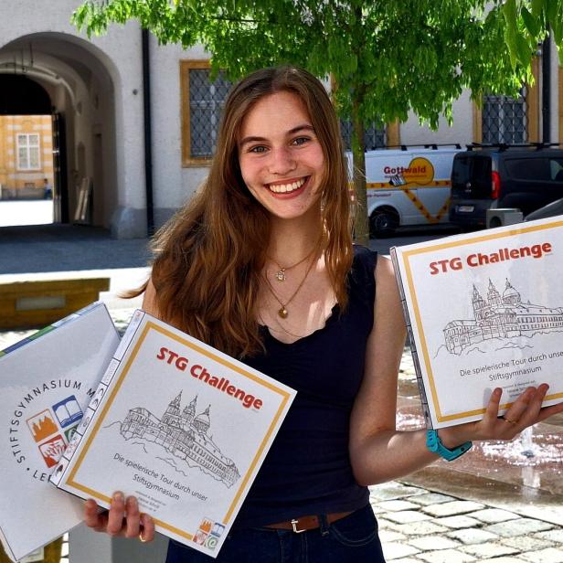 Junge Frau mit langen Haaren hält draußen mehrere Spielekartons mit der Aufschrift „STG Challenge“ und lächelt.