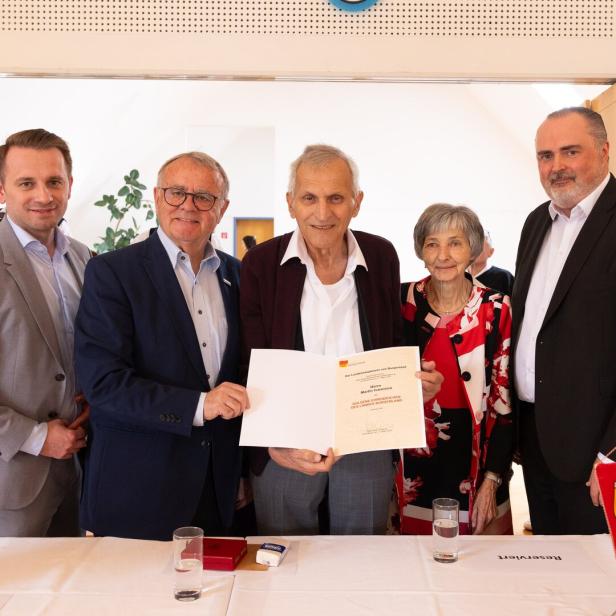Fünf Personen stehen zusammen, eine Person in der Mitte hält eine Urkunde, im Hintergrund spielen Musiker, auf dem Tisch stehen Wasser und Blumen.
