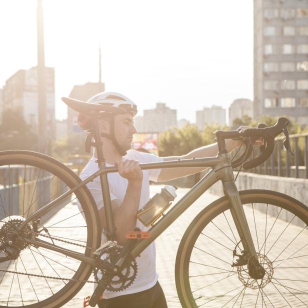 Ein Mann mit Fahrradhelm trägt ein Fahrrad auf der Schulter über eine Brücke in der Stadt bei Sonnenschein.