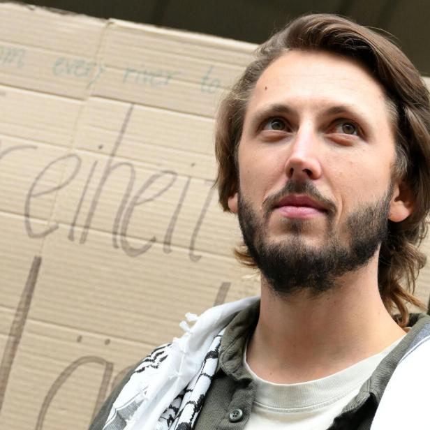 Ein Mann mit Bart steht vor einem Kartonschild mit der Aufschrift „Freiheit“ und trägt ein schwarz-weißes Tuch.