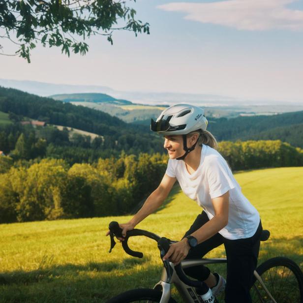 Eine Person mit Fahrradhelm fährt bei sonnigem Wetter durch eine grüne Hügellandschaft.