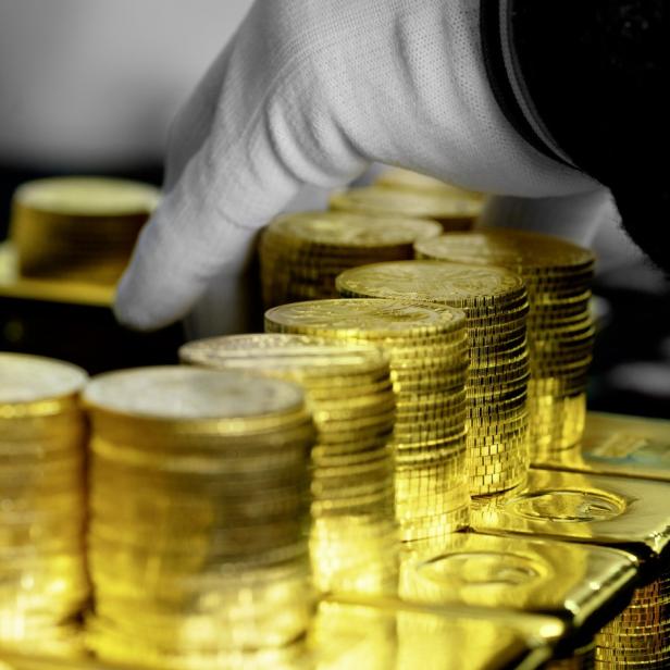 Gestapelte Goldmünzen und Goldbarren werden von einer Hand mit weißem Handschuh sortiert.