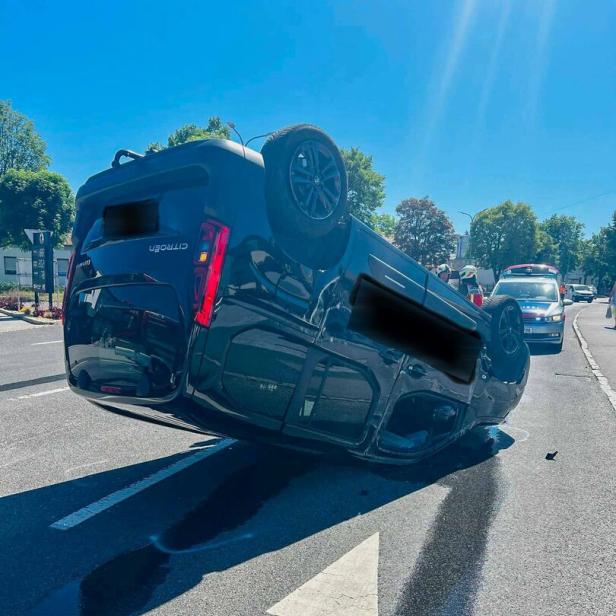 Ein Auto liegt auf dem Dach auf einer Straße, während Einsatzfahrzeuge und Personen im Hintergrund zu sehen sind.