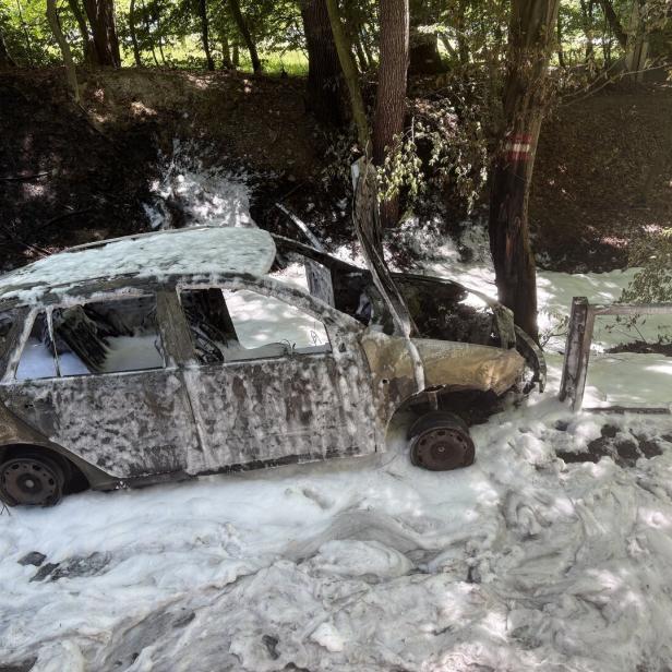 Ausgebranntes Auto mit Löschschaum bedeckt steht neben einem Baum und beschädigtem Geländer im Wald.