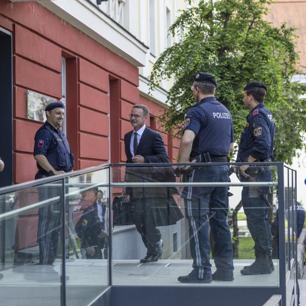 Ein Mann im Anzug geht an mehreren Polizisten vorbei, während im Hintergrund Kamerateams und Zuschauer stehen.