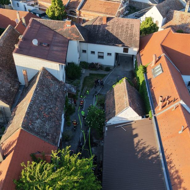 Mehrere Feuerwehrleute stehen in einem Innenhof zwischen Häusern mit roten Dächern und arbeiten mit Schläuchen und Leitern.