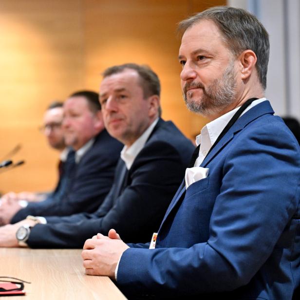 Vier Männer in Anzügen sitzen nebeneinander an einem Konferenztisch und hören aufmerksam zu.