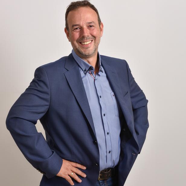 Ein Mann im blauen Anzug steht mit den Händen in die Hüften gestützt und lächelt freundlich vor hellem Hintergrund.