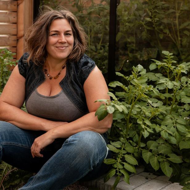 Eine Frau sitzt lächelnd in einem Gartenbeet, umgeben von üppigen grünen Pflanzen und trägt legere Kleidung.