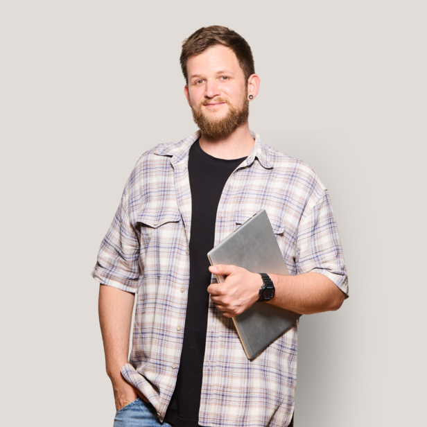 Ein junger Mann mit Bart und kariertem Hemd hält einen Laptop und lächelt vor hellem Hintergrund.