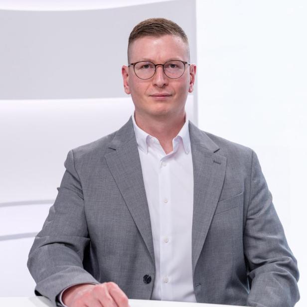 Mann mit Brille und kurzem Haar trägt grauen Anzug und weißes Hemd, sitzt an einem weißen Tisch vor hellem Hintergrund.