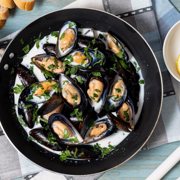 Miesmuscheln in cremiger Sauce mit Petersilie in einer Pfanne, dazu Zitronenscheiben und Baguette.