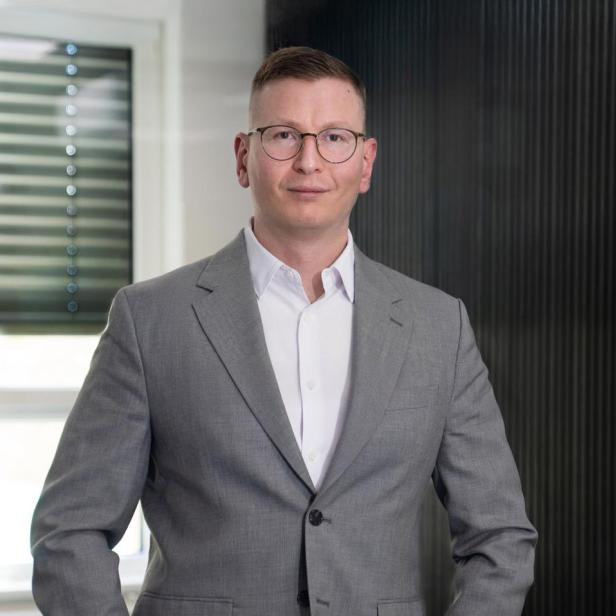 Ein Mann mit Brille und kurzem Haar trägt einen grauen Anzug und steht in einem modernen Büro mit Fenster im Hintergrund.