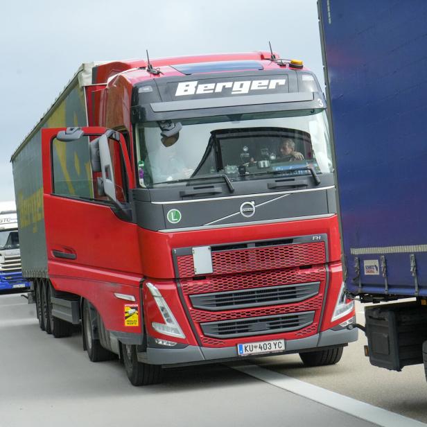 Ein roter Lkw fährt dicht neben einem blauen Lkw auf einer Autobahn, weitere Lkw sind im Hintergrund zu sehen.