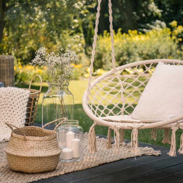 Eine gemütliche Terrasse mit Hängesessel, Pölster, Teppich, Körben, Glasvasen mit Blumen und Kerzen im Grünen.