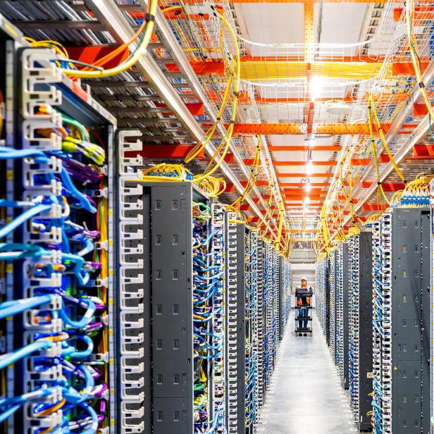 Reihen von Serverracks mit bunten Kabeln und einer Person, die einen Wagen durch einen Gang schiebt.
