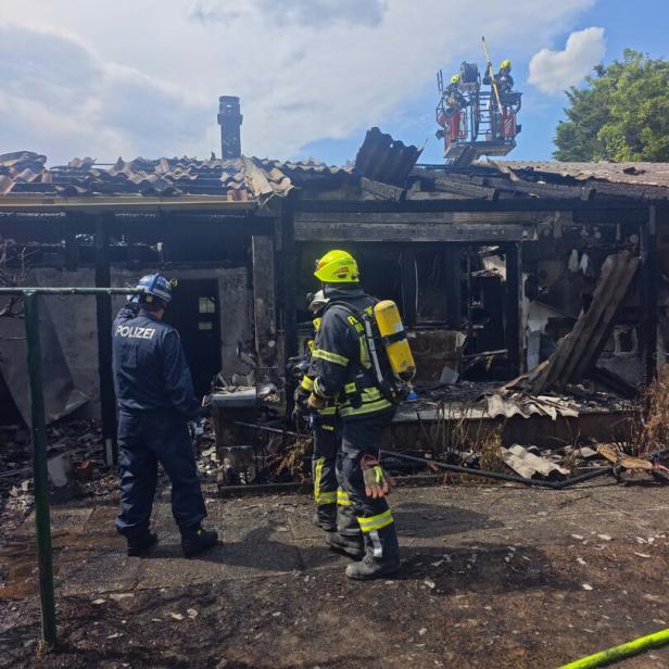 Zwei Feuerwehrleute und eine Polizistin stehen vor einem ausgebrannten Gebäude, während weitere Einsatzkräfte auf einer Drehleiter arbeiten.
