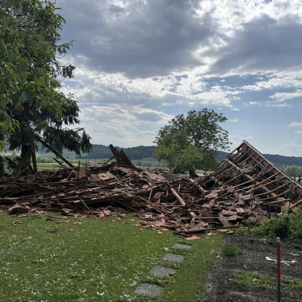 Ein eingestürztes Holzgebäude liegt in Trümmern auf einer Wiese, umgeben von Bäumen und ländlicher Landschaft.