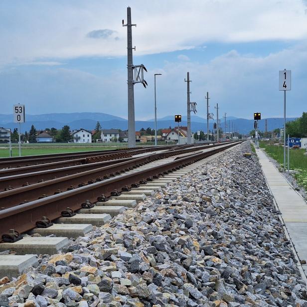Zwei Bahngleise verlaufen parallel neben einem Gehweg, im Hintergrund sind Häuser und Berge zu sehen.