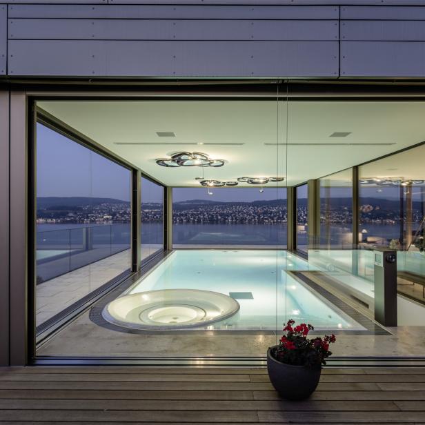 Modernes Wohnzimmer mit großem Innenpool, Whirlpool und Panoramablick auf einen See und eine Stadt im Hintergrund.