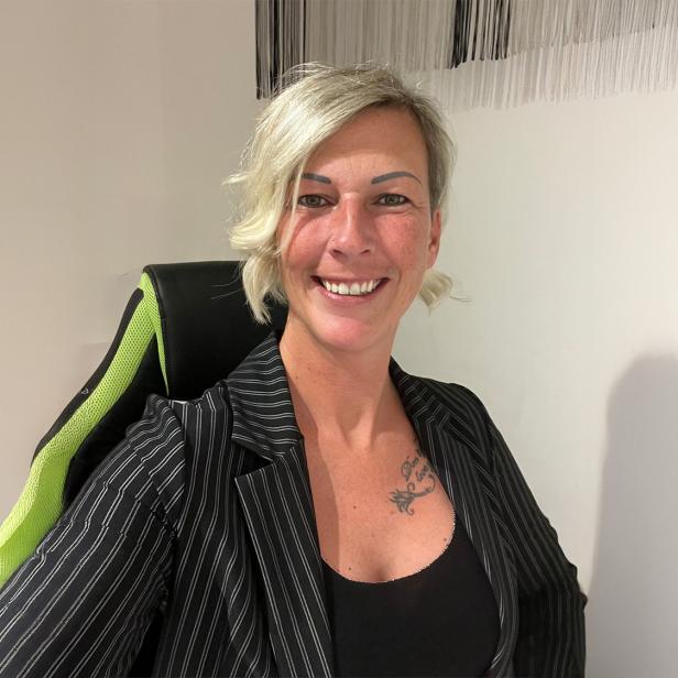 Eine lächelnde Person mit blondem, kurzem Haar sitzt auf einem Stuhl mit grünem Rückenlehnenbezug vor einer hellen Wand.