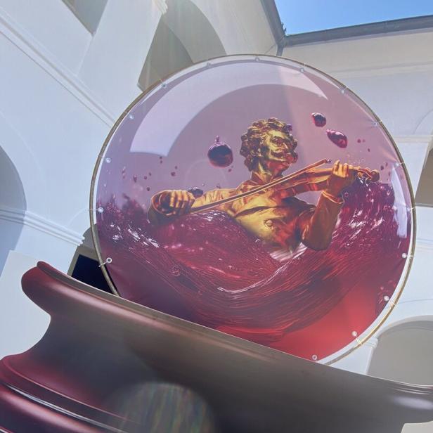 Eine goldene Figur spielt Geige in einer Glaskugel, die mit roter Flüssigkeit und Blasen gefüllt ist.