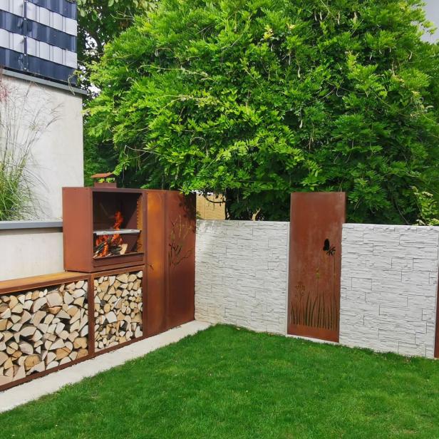 Ein Grill mit Holzlager aus rostigem Metall steht in einer modernen Gartenecke mit grüner Rasenfläche.