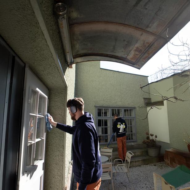 Zwei Personen reinigen Fenster in einem sonnigen Innenhof mit Gartenmöbeln und kahlen Ästen.