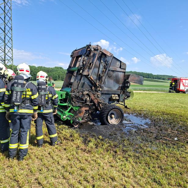 Mehrere Feuerwehrleute stehen vor einer ausgebrannten landwirtschaftlichen Maschine auf einem Feld, im Hintergrund ein Einsatzfahrzeug.