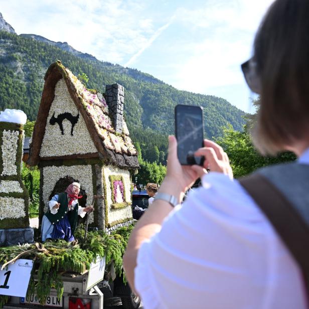 Eine Person fotografiert ein blumengeschmücktes Hexenhaus auf einem Wagen vor einer Bergkulisse.