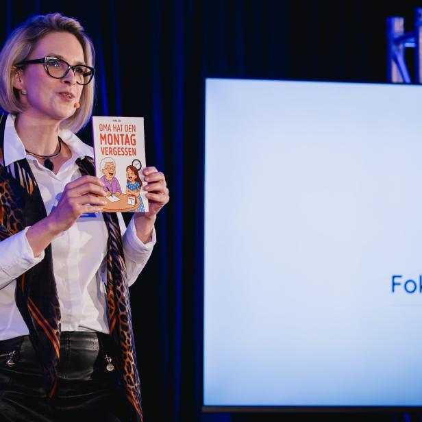 Eine Frau mit Brille hält ein Buch mit dem Titel „Oma hat den Montag vergessen“ vor einem Bildschirm mit dem Logo „FokusDemenz.at“.