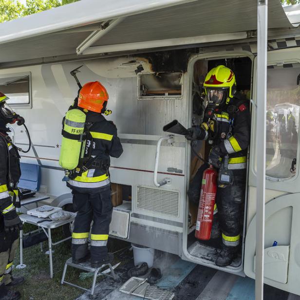 Drei Feuerwehrleute mit Atemschutz löschen einen Brand in einem Wohnmobil, das an der Tür und Wand beschädigt ist.