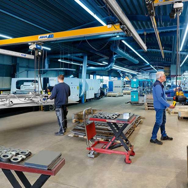 Zwei Männer bedienen einen Hallenkran in einer modernen, gut beleuchteten Produktionshalle.