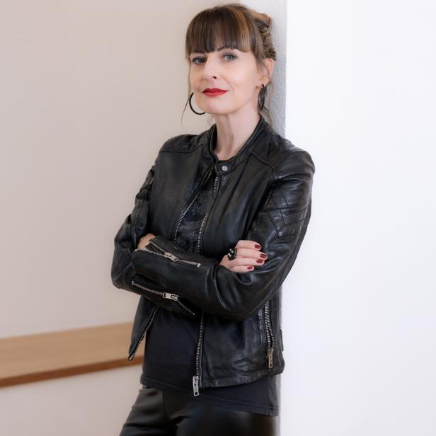 Frau mit dunkler Lederjacke und verschränkten Armen lehnt an einer weißen Wand.