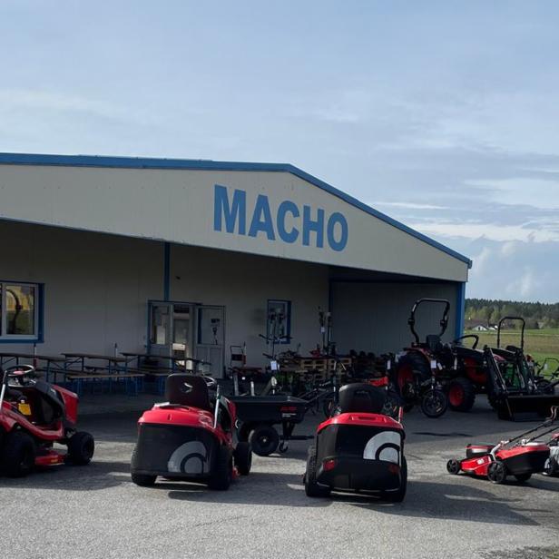 Vor einem Gebäude mit der Aufschrift „MACHO“ stehen mehrere rote Rasenmäher und landwirtschaftliche Maschinen.
