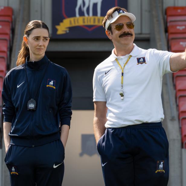 Ted Lasso zeigt auf dem Fußballfeld mit einer Frau im Trainingsanzug neben ihm.