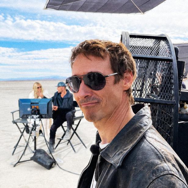Ein Mann mit Sonnenbrille und Lederjacke steht im Vordergrund, im Hintergrund sitzen zwei Personen an Monitoren in einer Wüstenlandschaft.