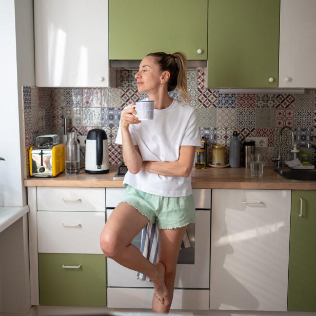 Satisfied happy woman standing alone in kitchen at cozy home atmosphere with cup of hot coffee.