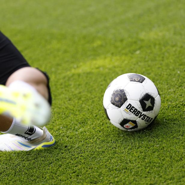 Ein Fußball liegt auf grünem Rasen, daneben ist ein Bein mit weißem Sportschuh in Bewegung zu sehen.
